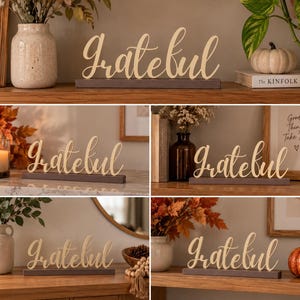 May include: A decorative tabletop sign featuring the word Grateful in a gold-colored cursive script font. The lettering is mounted on a rectangular dark gray base. The sign is shown in multiple settings with various seasonal home decor items.