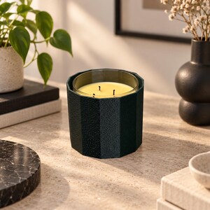 Puede incluir: Una vela de tres mechas en un recipiente de vidrio cil&iacute;ndrico de color verde oscuro, facetado y texturizado. La cera del interior es de color amarillo p&aacute;lido. El recipiente tiene una forma geom&eacute;trica distintiva de varios lados con acabado mate.
