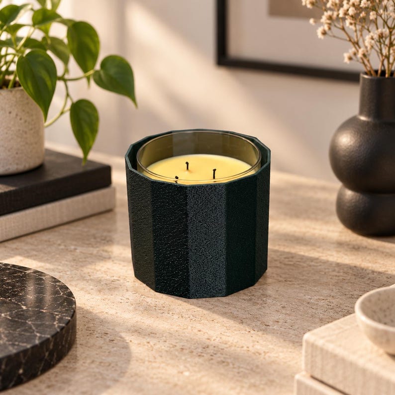Puede incluir: Una vela de tres mechas en un recipiente de vidrio cil&iacute;ndrico de color verde oscuro, facetado y texturizado. La cera del interior es de color amarillo p&aacute;lido. El recipiente tiene una forma geom&eacute;trica distintiva de varios lados con acabado mate.