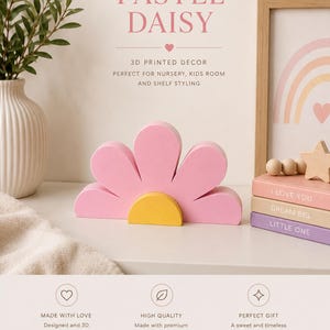 K&ouml;nnte beinhalten: Ein pastellrosa, g&auml;nsebl&uuml;mchenf&ouml;rmiges 3D-gedrucktes Dekorst&uuml;ck mit gelber Mitte. Das Bild zeigt auch eine Vase mit Gr&uuml;n, einen gerahmten Regenbogen-Druck und gestapelte B&uuml;cher mit den S&auml;tzen "I LOVE YOU", "DREAM BIG" und "LITTLE ONE". Der Text lautet "PASTEL DAISY".