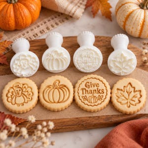 May include: Four white plastic cookie stamps with handles and four round cookies featuring embossed designs of a turkey, a pumpkin, the text Give Thanks with a small pumpkin, and a maple leaf, all arranged on a wooden board.