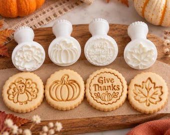 Puede incluir: Cuatro sellos de plástico blanco para galletas con mangos y cuatro galletas redondas con diseños en relieve de un pavo, una calabaza, el texto Give Thanks con una calabaza pequeña y una hoja de arce sobre una tabla.