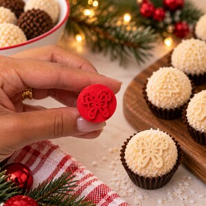 May include: A small, circular red plastic stamp featuring an embossed butterfly design. The stamp is held up to display the intricate butterfly pattern, which matches the white butterfly-shaped decoration on a round, coconut-covered confection.