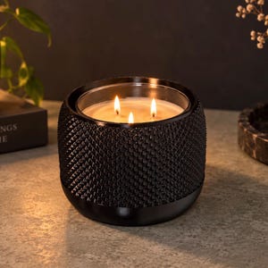 May include: A three-wick candle in a cylindrical black container with a textured, diamond-patterned exterior. The wax is light-colored and the three wicks are lit, producing a warm glow that illuminates the interior of the glass vessel.