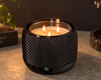 May include: A three-wick candle in a cylindrical black container with a textured, diamond-patterned exterior. The wax is light-coloured and the three wicks are lit, producing a warm glow that illuminates the interior of the glass vessel.