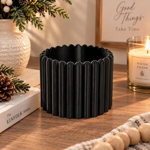 May include: A matte black cylindrical planter featuring a vertical fluted or ribbed texture along the exterior. The container has a scalloped top edge and a hollow interior, designed with a consistent, repeating pattern of rounded vertical columns.