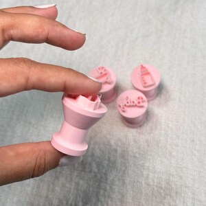 May include: A set of four light pink plastic fondant stamps featuring baby-themed designs. One stamp is held up to show the cylindrical handle and the raised impression surface, while three others sit nearby with icons of a bottle and the word baby.