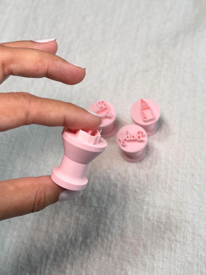 May include: A set of four light pink plastic fondant stamps featuring baby-themed designs. One stamp is held up to show the cylindrical handle and the raised impression surface, while three others sit nearby with icons of a bottle and the word baby.
