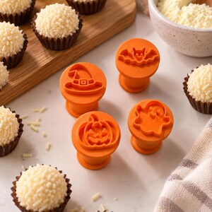 May include: Four orange plastic stamps for decorating food, each featuring a distinct Halloween-themed design: a witch hat, a bat, a pumpkin, and a ghost. These circular tools are arranged on a surface alongside several small white treats.