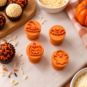 May include: Four orange plastic cookie stamps featuring raised designs of a witch hat, a bat, a jack-o'-lantern, and a ghost. Each stamp has a cylindrical handle and a flat base for pressing patterns into dough, arranged on a light surface.