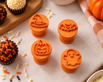 May include: Four orange plastic cookie stamps featuring raised designs of a witch hat, a bat, a jack-o'-lantern, and a ghost. Each stamp has a cylindrical handle and a flat base for pressing patterns into dough, arranged on a light surface.