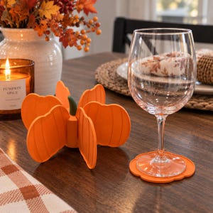 May include: A bright orange silicone pumpkin-shaped coaster set that can be assembled into a 3D pumpkin figure. The set includes a circular coaster base for a clear glass wine goblet, featuring a smooth surface and a small green stem component.
