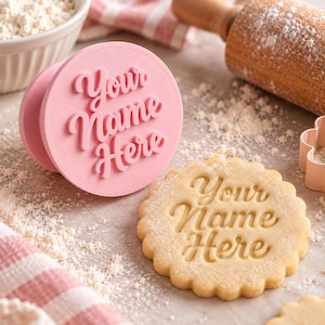 May include: A round pink cookie stamp featuring the raised text Your Name Here in a cursive font, positioned next to a scalloped-edge cookie with the same text impressed into the surface, surrounded by scattered flour and baking tools.