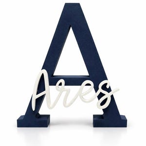 May include: A navy blue wooden letter "A" with the name "Ares" in white script. The letter is freestanding and has a textured surface. The name is in a cursive font.