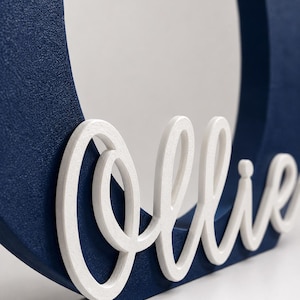 Puede incluir: Un marco de madera azul marino con el nombre "Ollie" en escritura blanca. El marco tiene una abertura circular y una superficie texturizada. El nombre est&aacute; en una fuente cursiva.