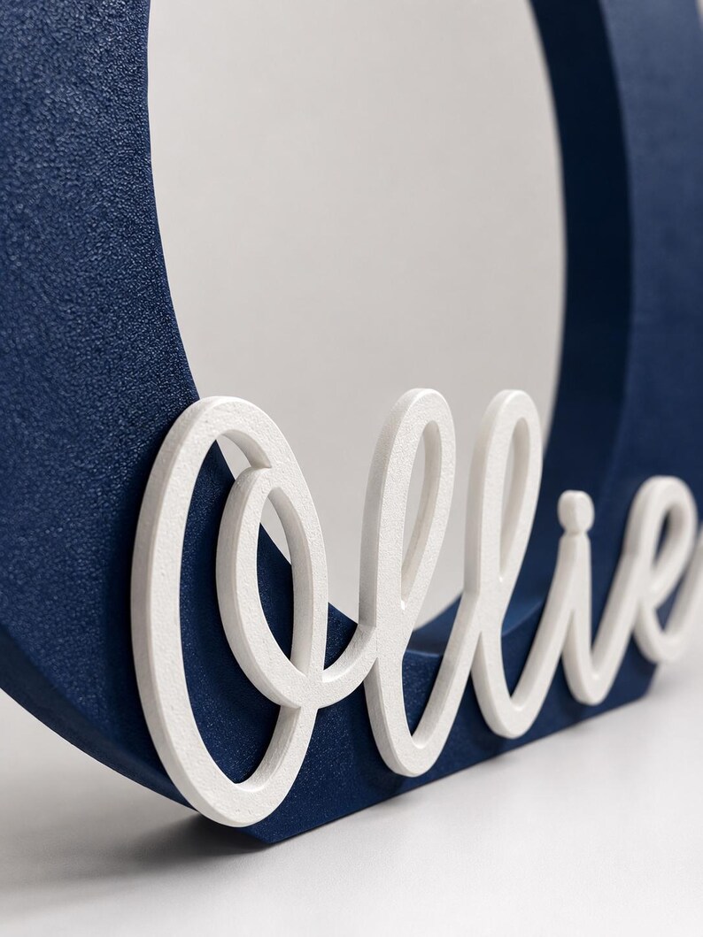 Puede incluir: Un marco de madera azul marino con el nombre "Ollie" en escritura blanca. El marco tiene una abertura circular y una superficie texturizada. El nombre est&aacute; en una fuente cursiva.