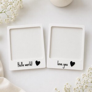 May include: Two white square photo frames with a classic instant camera border design. The left frame features the black text Hello world! and a black heart. The right frame features the black text love you and a black heart.