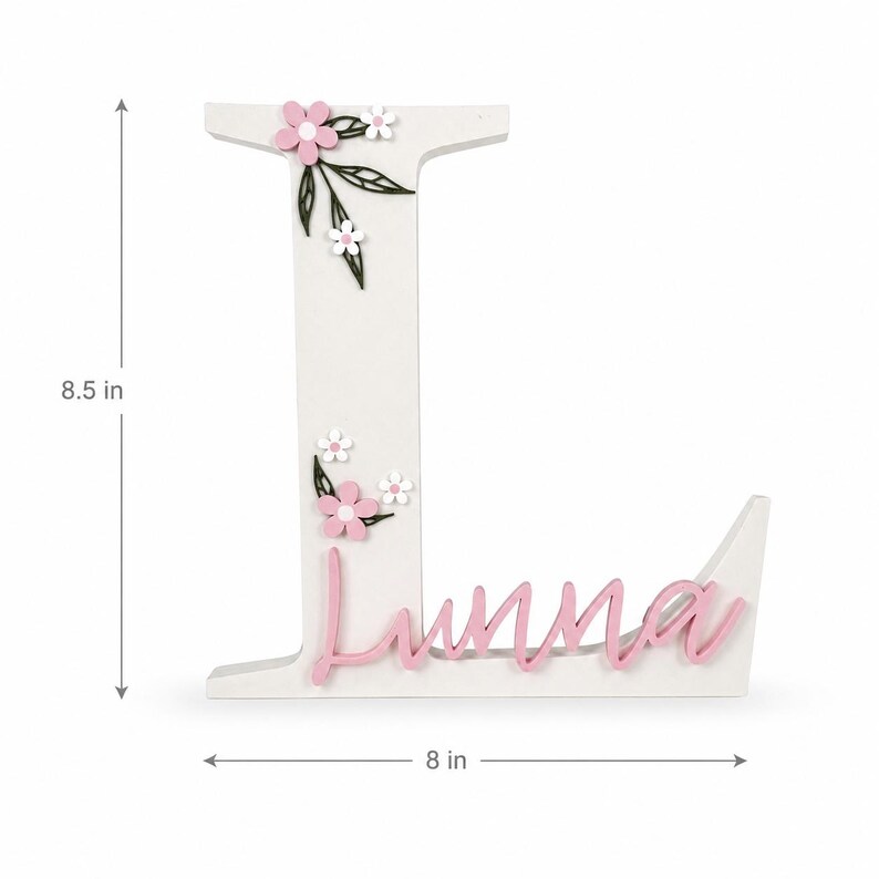 May include: White wooden letter "L" decorated with pink flowers, white flowers, and green leaves. The name "Luna" is written in pink script. The letter is 21.6 cm tall and 20.3 cm wide.