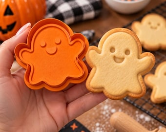 Puede incluir: Un cortador de galletas de plástico naranja con forma de fantasma junto a una galleta horneada de color tostado claro con la misma forma. El cortador presenta el contorno simple de un fantasma con dos ojos circulares y una boca curva.