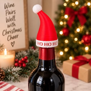 May include: A red and white Santa hat bottle topper featuring a white pom-pom at the tip and a white band around the base with the text HO HO HO printed in red. The hat is positioned on the neck of a dark glass bottle.