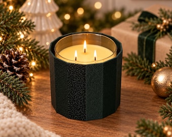 Puede incluir: Una vela de tres mechas en un recipiente cilíndrico de color verde oscuro, facetado y texturizado. La cera es de color crema pálido y las tres mechas están encendidas, produciendo llamas pequeñas y brillantes. El recipiente tiene una forma geométrica.