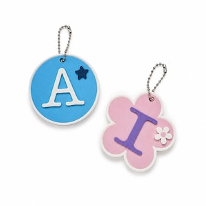 Puede incluir: Dos llaveros decorativos. Uno es un c&iacute;rculo azul con la letra blanca "A" y una estrella azul. El otro es una forma de flor rosa con la letra morada "I" y una flor blanca. Ambos tienen fijaciones de cadena de bolas plateadas.