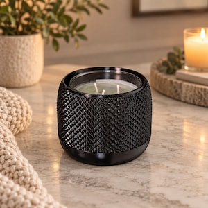 May include: A black candle in a cylindrical container with a textured, diamond-patterned exterior. The candle features three white wicks centered in light-colored wax. The container has a smooth, solid black base and a wide, open top rim.