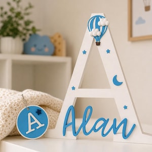 Puede incluir: Una letra "A" de madera blanca con detalles azules, incluyendo un globo aerost&aacute;tico, estrellas y el nombre "Alan" en cursiva. Una etiqueta redonda azul con la letra "A" y una estrella cuelga cerca. Decoraci&oacute;n de guarder&iacute;a.