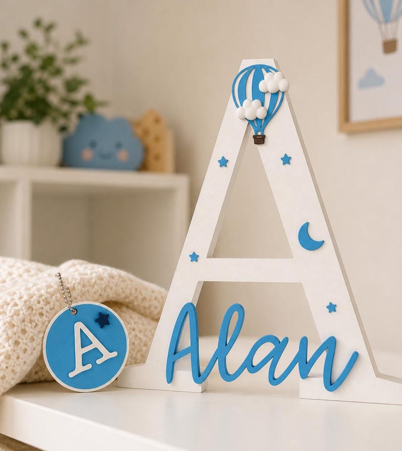 Puede incluir: Una letra "A" de madera blanca con detalles azules, incluyendo un globo aerost&aacute;tico, estrellas y el nombre "Alan" en cursiva. Una etiqueta redonda azul con la letra "A" y una estrella cuelga cerca. Decoraci&oacute;n de guarder&iacute;a.