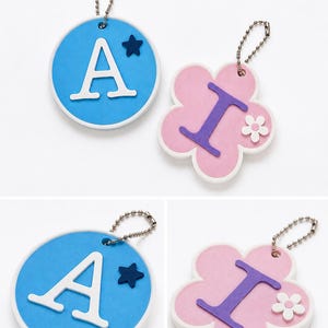 Puede incluir: Dos llaveros: uno redondo, azul con una "A" blanca y una estrella azul; el otro, con forma de flor rosa con una "I" morada y una flor blanca. Ambos tienen enganches de cadena plateada.