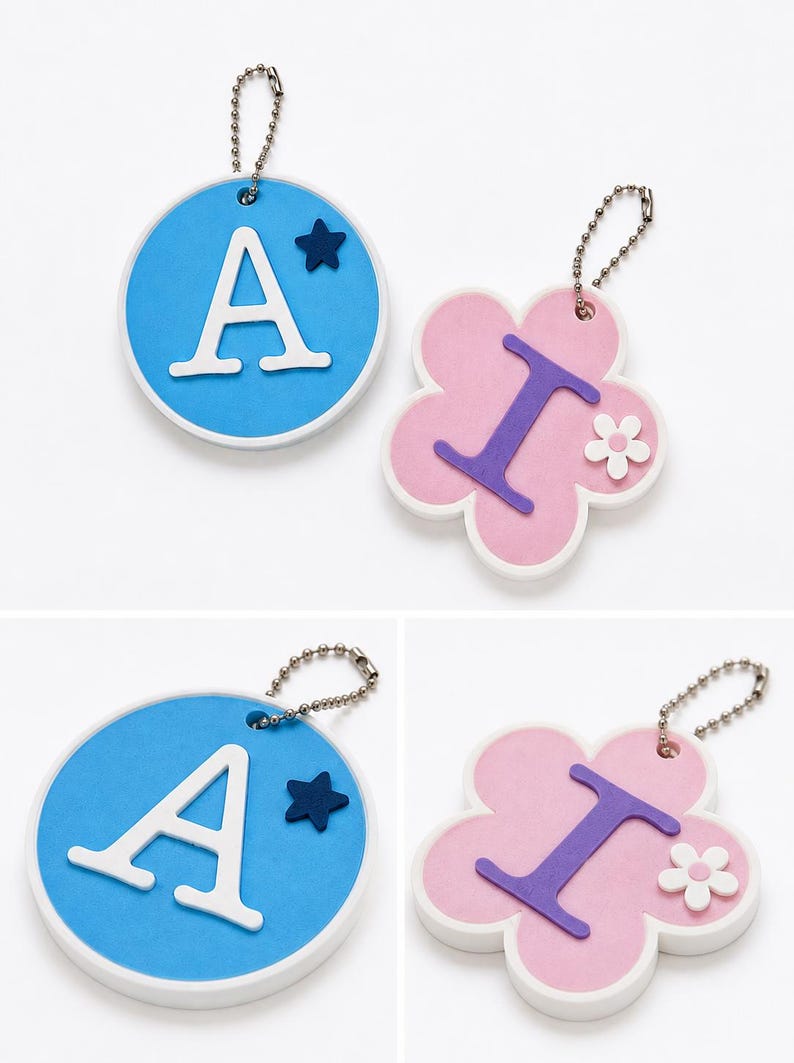 Puede incluir: Dos llaveros: uno redondo, azul con una "A" blanca y una estrella azul; el otro, con forma de flor rosa con una "I" morada y una flor blanca. Ambos tienen enganches de cadena plateada.