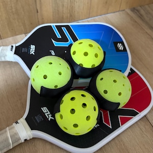 Puede incluir: Dos palas de pickleball con mangos blancos y empu&ntilde;aduras negras, una con detalles en azul y negro y la otra en rojo y negro. Cuatro pelotas de pickleball de color amarillo ne&oacute;n perforadas est&aacute;n dispuestas sobre las palas, sujetas por clips de pl&aacute;stico negro.