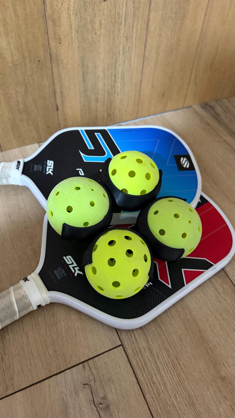 Puede incluir: Dos palas de pickleball con mangos blancos y empu&ntilde;aduras negras, una con detalles en azul y negro y la otra en rojo y negro. Cuatro pelotas de pickleball de color amarillo ne&oacute;n perforadas est&aacute;n dispuestas sobre las palas, sujetas por clips de pl&aacute;stico negro.