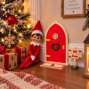 May include: A red elf doll with a white collar and hat sits beside a miniature arched red door with a green wreath, a small yellow handle, and a welcome mat featuring a tree. A wooden sign nearby reads Elf Door in red letters.