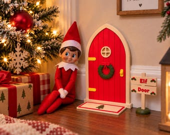 May include: A red elf doll with a white collar and hat sits beside a miniature arched red door with a green wreath, a small yellow handle, and a welcome mat featuring a tree. A wooden sign nearby reads Elf Door in red letters.