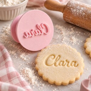 May include: A round pink plastic cookie stamp featuring the name Clara in raised cursive script, positioned next to a matching scalloped-edge cookie embossed with the same name, with a light dusting of flour visible on the surface.