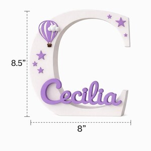 Może przedstawiać: Biała drewniana litera "C" z imieniem "Cecilia" w fioletowym skrypcie. Litera ma 21,6 cm wysokości i 20,3 cm szerokości. Zawiera fioletowy balon na ogrzane powietrze, chmury i gwiazdy. Element dekoracyjny do pokoju dziecięcego.