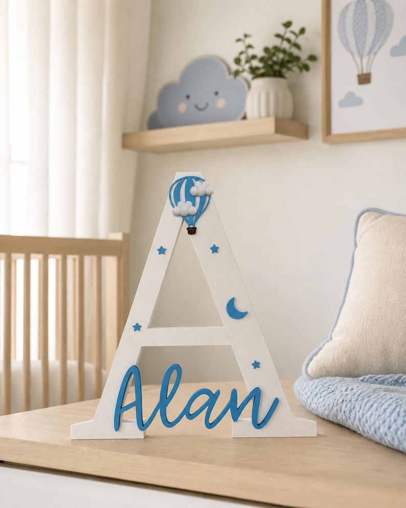 May include: A white wooden letter "A" with blue accents, featuring a hot air balloon and stars. The name "Alan" is written in blue script. Decorative item for a nursery or child's room.