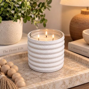 May include: A three-wick candle in a white cylindrical vessel with a textured, ribbed exterior. The candle features a clear glass insert holding white wax with three lit wicks, creating a warm glow. The vessel has a matte finish and horizontal ridges.