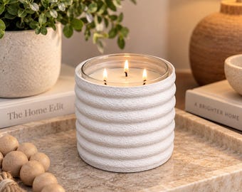 May include: A three-wick candle in a white cylindrical vessel with a textured, ribbed exterior. The candle features a clear glass insert holding white wax with three lit wicks, creating a warm glow. The vessel has a matte finish and horizontal ridges.