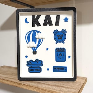 May include: A square shadow box frame with a black border containing a white background. The name KAI is displayed in black glitter letters. Blue icons include a hot air balloon with white clouds, an alarm clock showing 10:25 AM, a calendar showing February 27, a baby on a scale labeled 6LB 11OZ, and a ruler labeled 19.7INCHES.