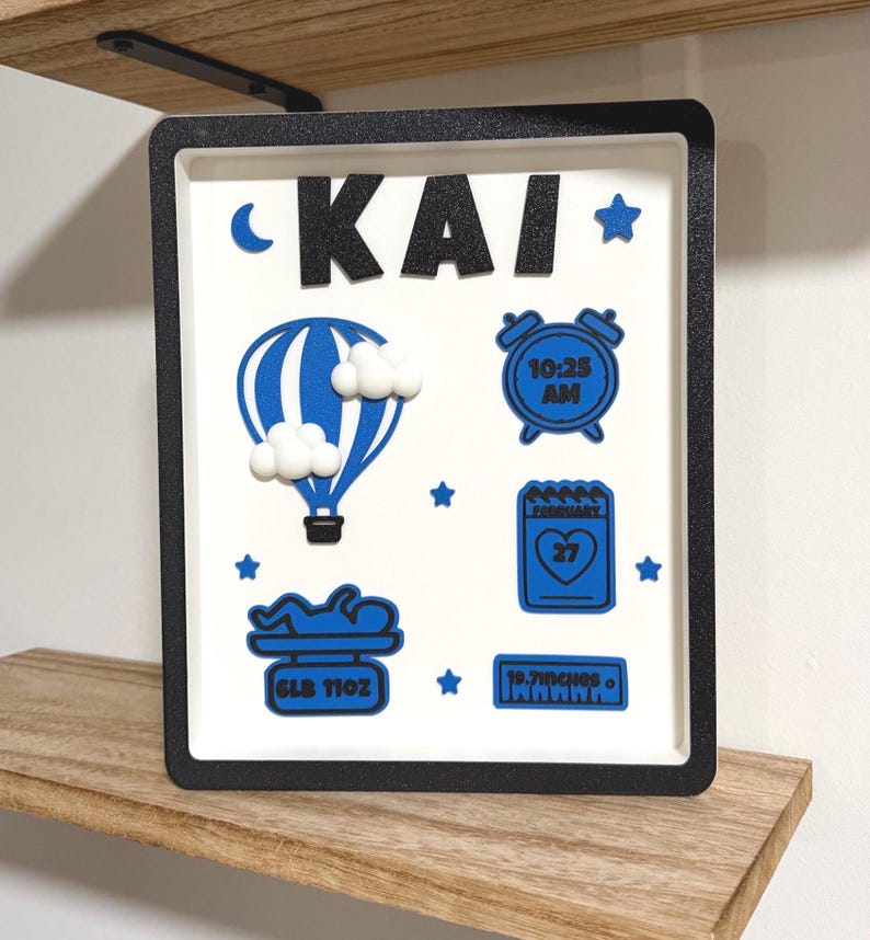 May include: A square shadow box frame with a black border containing a white background. The name KAI is displayed in black glitter letters. Blue icons include a hot air balloon with white clouds, an alarm clock showing 10:25 AM, a calendar showing February 27, a baby on a scale labeled 6LB 11OZ, and a ruler labeled 19.7INCHES.