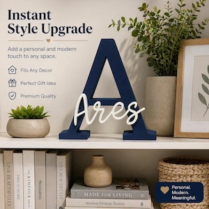 May include: A navy blue wooden letter "A" with the name "Ares" in white script. The decorative piece is displayed on a white shelf with books, a small plant, and other home decor items. The text "Instant Style Upgrade" is visible.