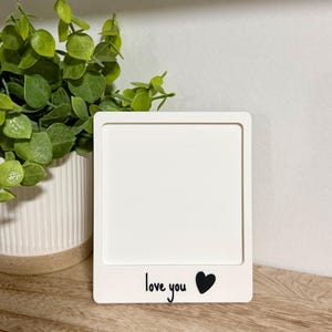 May include: A square white photo frame with a flat border. The bottom section of the frame features the black printed text "love you" followed by a solid black heart icon. The frame is designed to hold a square image in the center area.