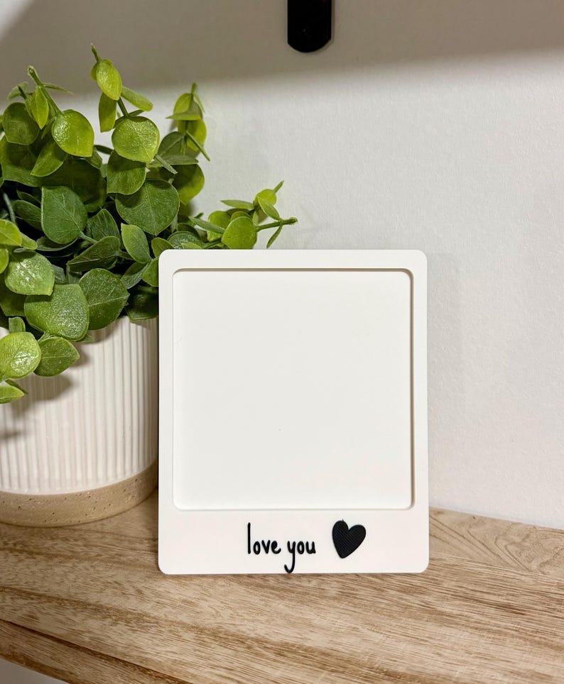 May include: A square white photo frame with a flat border. The bottom section of the frame features the black printed text "love you" followed by a solid black heart icon. The frame is designed to hold a square image in the center area.