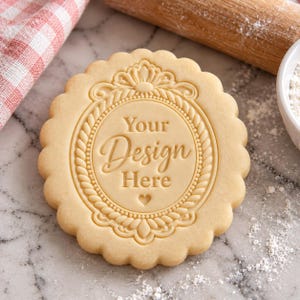 May include: A round, scalloped-edge cookie dough stamp featuring an ornate, rope-style circular frame with a decorative crest at the top. The center is embossed with the text Your Design Here and a small heart icon at the bottom.