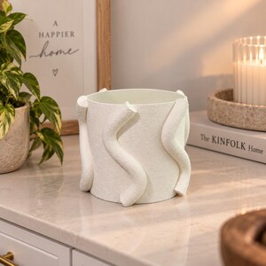 May include: A cylindrical white planter featuring a textured surface with three raised, vertical, wavy, snake-like decorative elements wrapping around the exterior. The planter has a smooth interior and a matte finish on the outer surface.