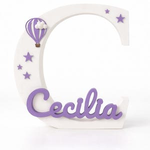 May include: A white wooden letter "C" with the name "Cecilia" in purple script. The letter is decorated with purple stars and a purple hot air balloon with a white cloud.