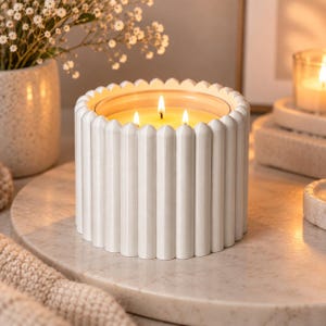 May include: A white cylindrical candle holder with a vertical ribbed texture, containing a three-wick candle with a glowing yellow flame. The candle is positioned on a circular stone surface, emitting a warm light from the burning wicks.