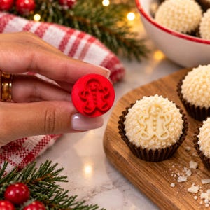 May include: A small, circular red plastic stamp featuring a raised gingerbread man design. The stamp is held up to show the embossed detail, which matches the gingerbread man shape imprinted on the white, textured round confection sitting nearby.